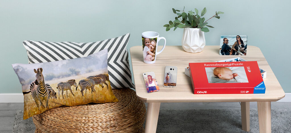Ein gemütlicher Tisch mit CEWE Produkten: einer personalisierten Tasse, einem Puzzle mit Babyfoto, sowie zwei Fotos auf dem Tisch und einem Kissen mit Zebras.