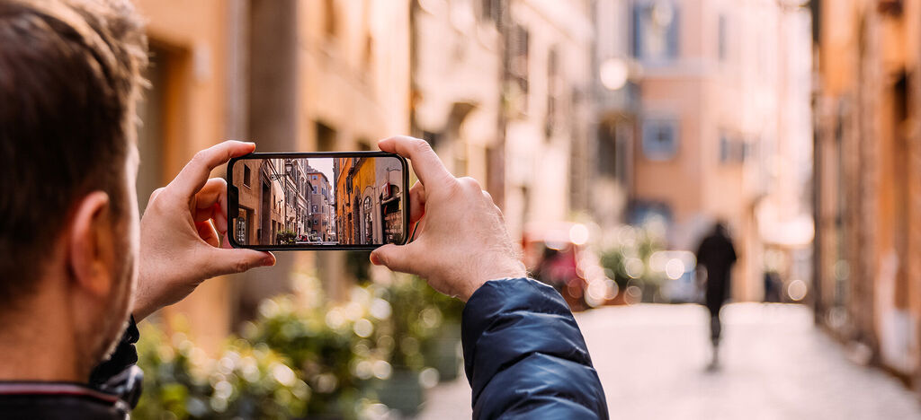 Ein Mann fotografiert eine belebte Gasse mit seinem Smartphone in einer Stadt.