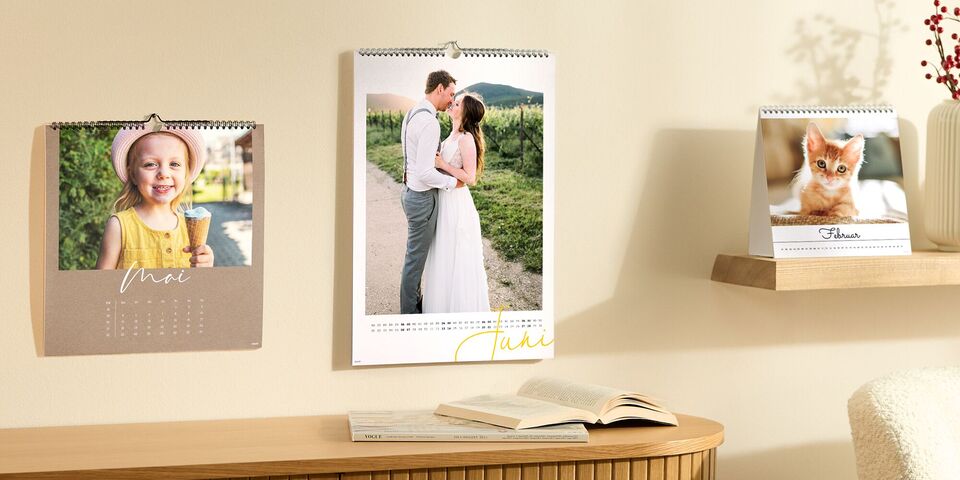 Ein Bild mit Fotokalendern von CEWE. 2 Kalender an einer Wand, mit Bildern eines Kinds und einem Paar. Ein Kalender stehend auf einem Regal.