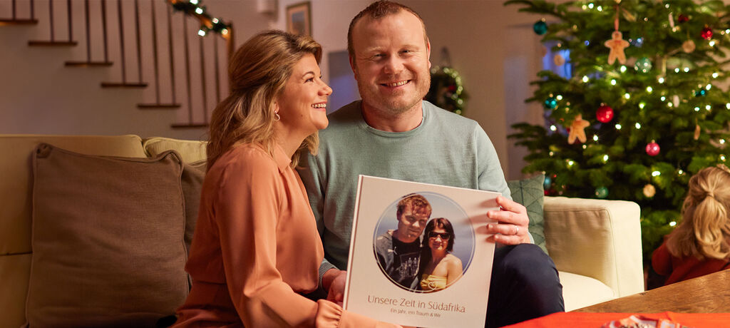 Ein Paar sitzt gemütlich auf dem Sofa und hält ein CEWE FOTOBUCH mit dem Titel 'Unsere Zeit in Südafrika' in den Händen. Der Weihnachtsbaum im Hintergrund verleiht dem Raum eine festliche Stimmung.