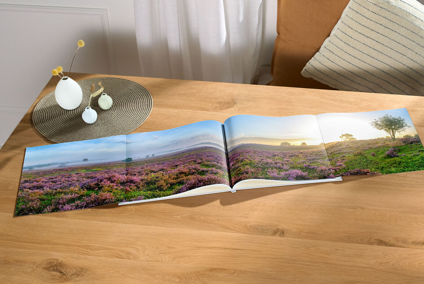 Ein geöffnetes CEWE FOTOBUCH liegt auf einem Holztisch mit einer blühenden Landschaft auf der linken Seite und einem gemütlichen Wohnzimmer im Hintergrund.