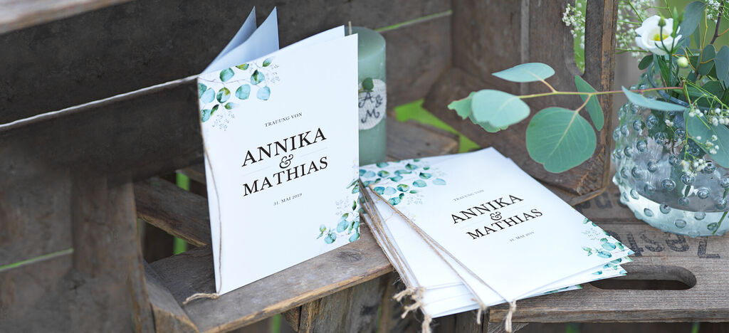 Zwei personalisierte CEWE Grußkarten mit dem Schriftzug "ANNIKA & MATHIAS" liegen auf einer Holzbank, umgeben von Pflanzen.