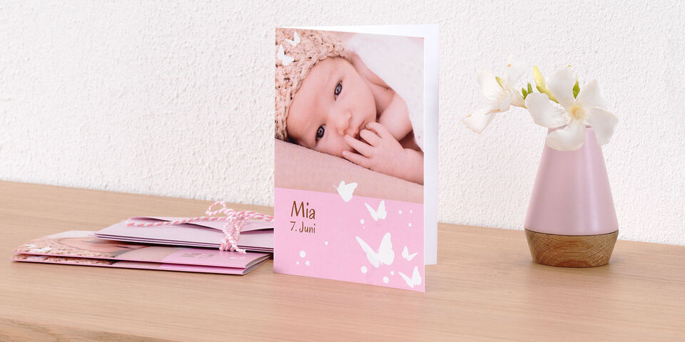 Eine zarte, rosa Grußkarte mit einem Foto eines schlafenden Babys. Auf der Karte steht "Mia 7. Juni" in einer liebevollen Schrift. Daneben steht eine dekorative Vase mit Blumen.