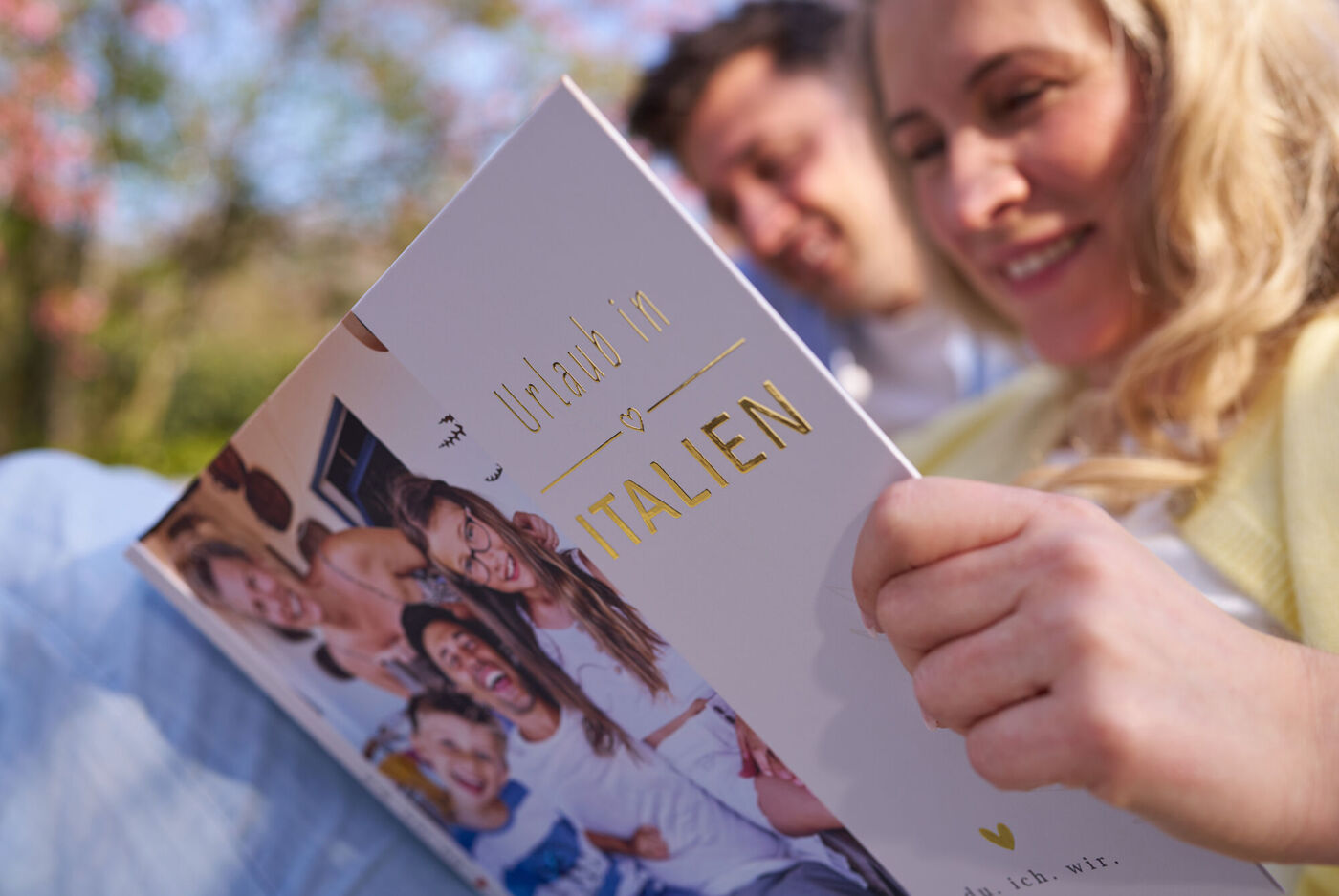Zwei Personen sitzen im Freien und betrachten gemeinsam ein Fotobuch mit Familienfoto auf dem Cover.