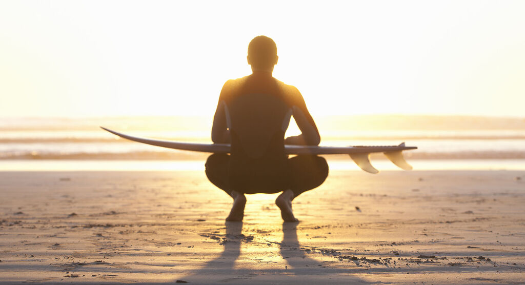 Surfer in der Hocke am Strand bei Sonnenuntergang, mit Surfbrett in der Hand, entspannte Atmosphäre