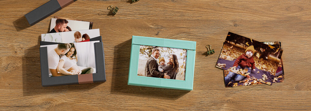 Drei verschiedene Fotoprodukte von CEWE auf einem Holztisch: Ein quadratischer Karton mit einem Bild von einer Familie auf der Vorderseite, ein Stapel von Fotos und eine praktische Box mit Bildern. Alle Produkte strahlen Freude und schöne Erinnerungen au