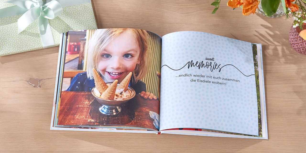 Ein offenes CEWE FOTOBUCH mit einem Eisbecher auf einer Holztafel. Die Seite zeigt den Text "endgültig weder die süßen Erinnerungen!"