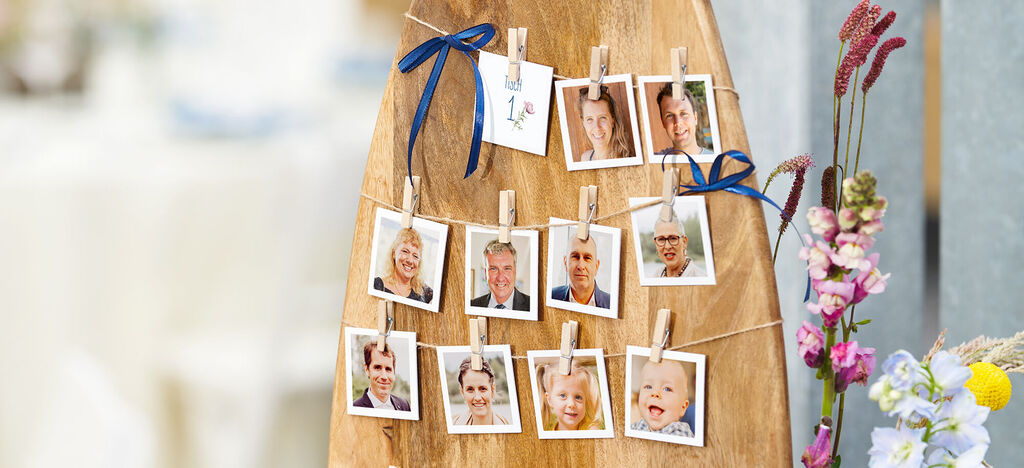 Eine dekorative Wand zeigt eine Auswahl von Fotos mit kleinen Holzklammern, auf einem Holzbrett, umgeben von zarten Blumen.
