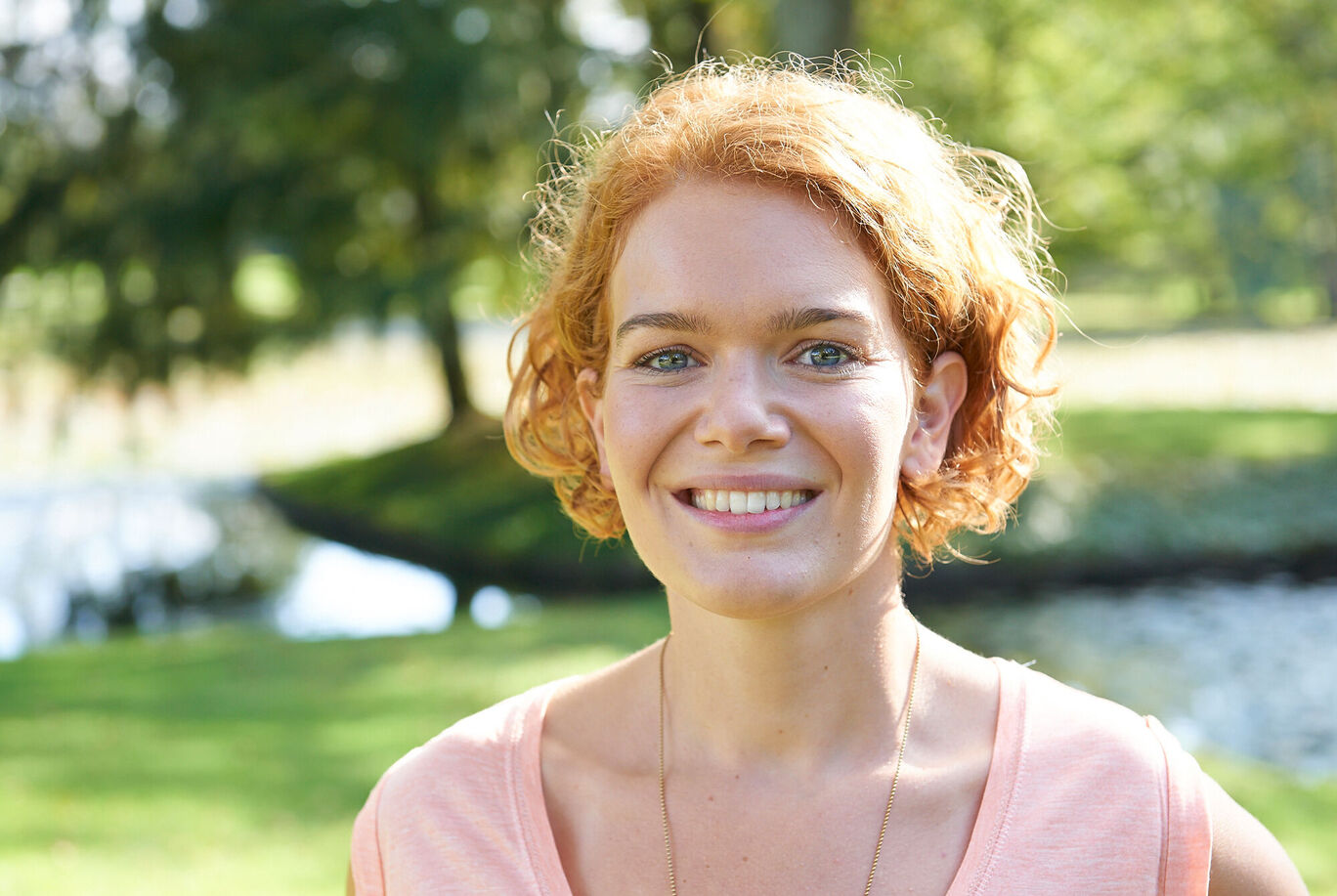 Eine Person mit Locken steht im Freien, umgeben von grünem Gras und Bäumen.