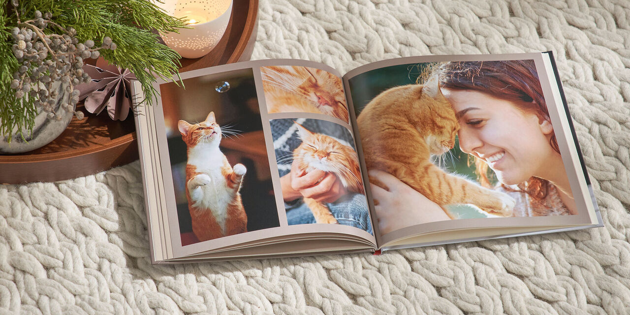 Ein aufgeschlagenes CEWE FOTOBUCH im quadratischen Format liegt auf einer weißen Decke. Auf der Doppelseite sind Fotos einer roten Katze zu sehen. Links neben dem Fotobuch liegt ein Tablett mit weihnachtlicher Deko und Kerzen.