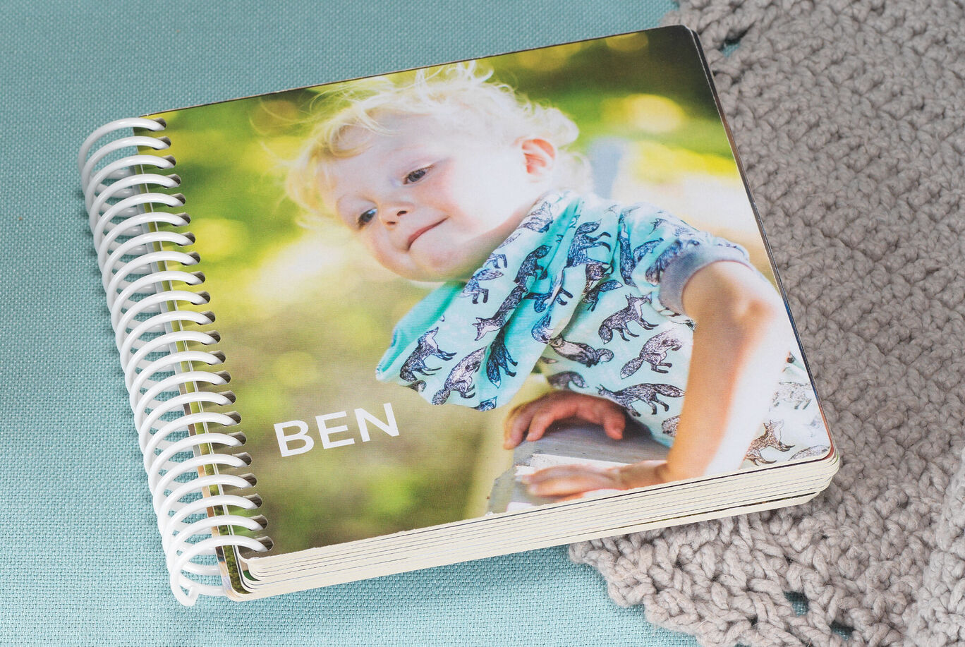 Ein liebevoll gestaltetes CEWE FOTOBUCH mit einem Jungen auf der Vorderseite. Der Junge trägt einen bunten Schal und ist in der Natur. Der Name „BEN“ ist auf dem Fotobuch abgebildet.