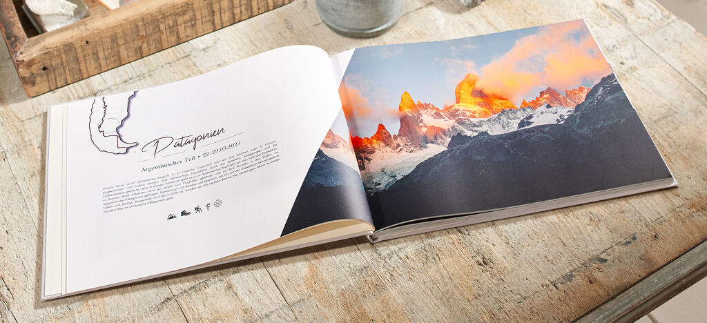 Ein offenes CEWE FOTOBUCH mit einem Bild der patagonischen Berge bei Sonnenuntergang auf einem Holztisch