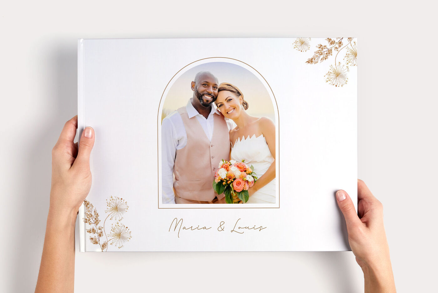 Zwei Hände halten ein CEWE FOTOBUCH XXL Panorama mit dem Foto eines Hochzeitspaarfotos in Bogenrahmen und Schriftzug "Maren & Lucas"