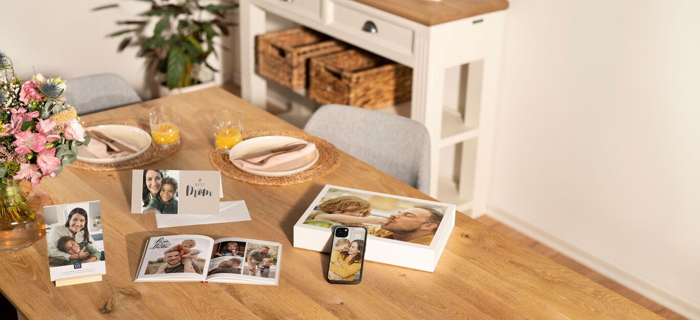 Der Tisch ist schön gedeckt mit einer CEWE Grußkarte, einem CEWE FOTOBUCH, einer persönlichen Fotokarte und einer Tasse. In der Hintergrund sind eine Pflanze und ein Kästchen zu sehen.
