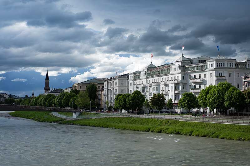 Blick auf eine malerische Uferpromenade mit eleganten Gebäuden und grünen Bäumen entlang einer ruhigen Gewässeroberfläche