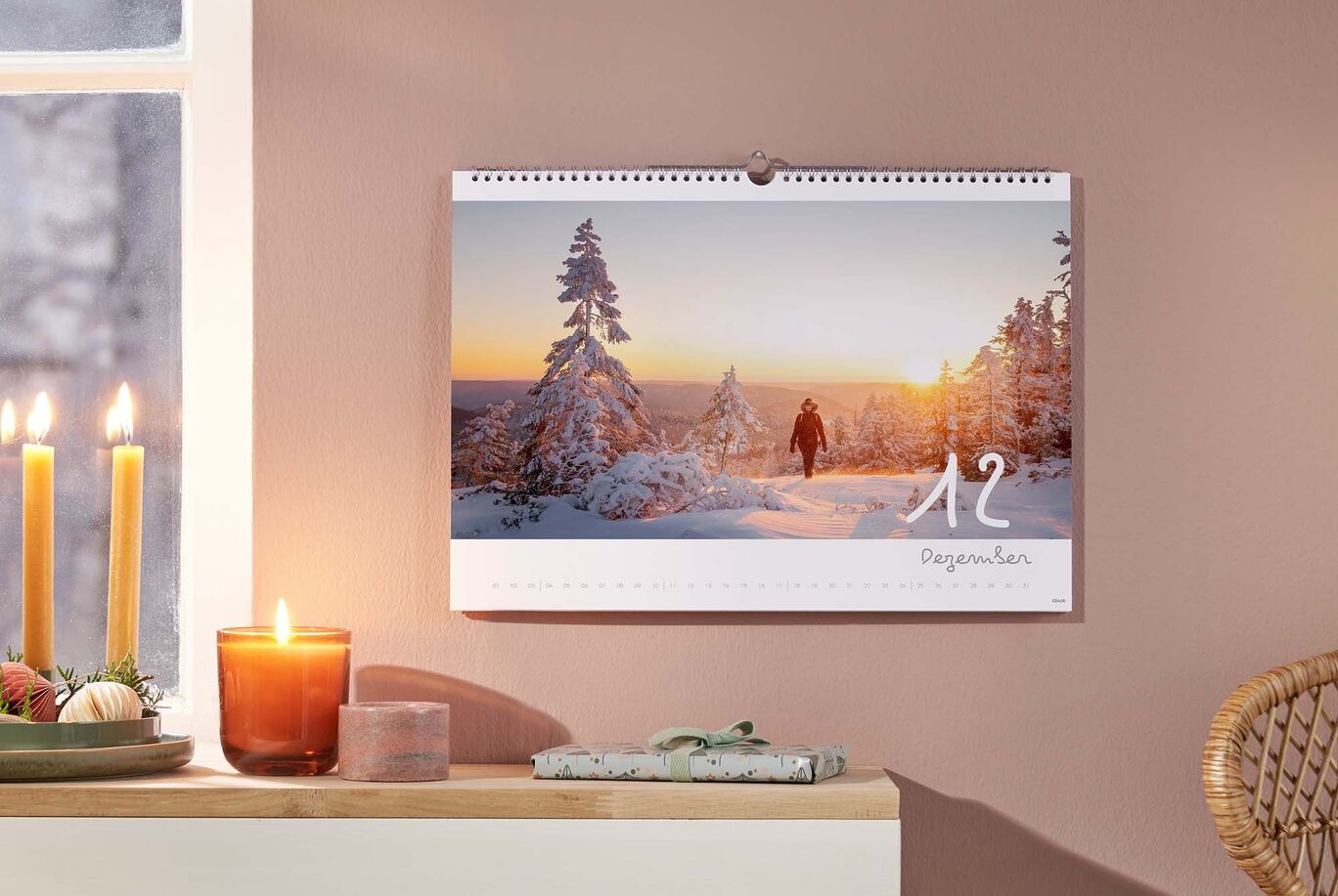 Ein wunderschöner Wandkalender mit winterlichen Landschaftsaufnahmen hängt an einer Wand. Die Szene zeigt schneebedeckte Bäume und einen Spaziergänger bei Sonnenuntergang.