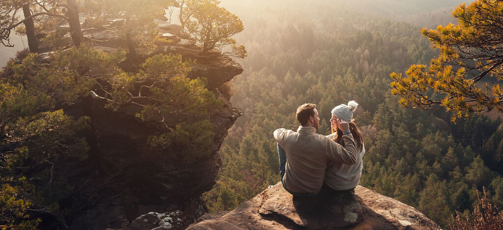 Ein Paar sitzt eng umschlungen auf einem Felsen mit Blick auf eine bewaldete Landschaft im Sonnenuntergang.