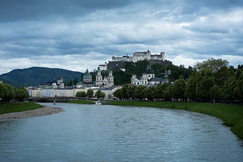 Das Bild zeigt die beeindruckende Festung Hohensalzburg in Salzburg, Österreich, die majestätisch auf einem Hügel thront. Im Vordergrund fließt ein ruhiger Fluss, umgeben von grünem Ufer.