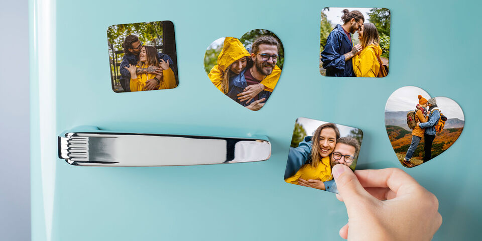 Ein Hand greift nach einem Herzmotiv-Foto auf einem Kühlschrank, umgeben von weiteren Herzfotos und einem Silbergriff.