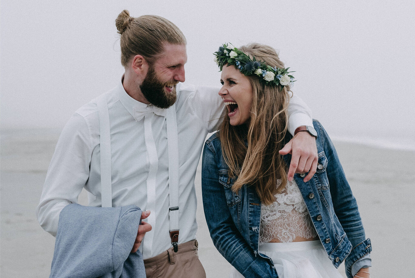 Ein Paar in Hochzeitskleidung am Strand, umarmt mit Freude, die Braut trägt einen Blumenkranz.