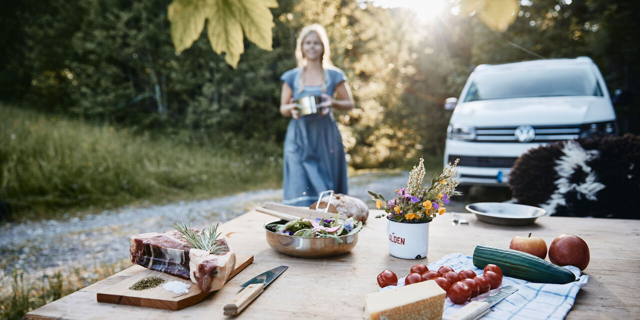 Gedeckter Tisch mit Grillfleisch, Käse und Gemüse. Eine Frau nähert sich von hinten, um zusätzliche Zutaten auf den Tisch zu stellen.