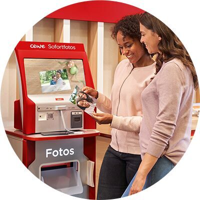 Zwei Frauen schauen sich Fotos an, die sie am CEWE Sofortfotoautomaten drucken lassen.