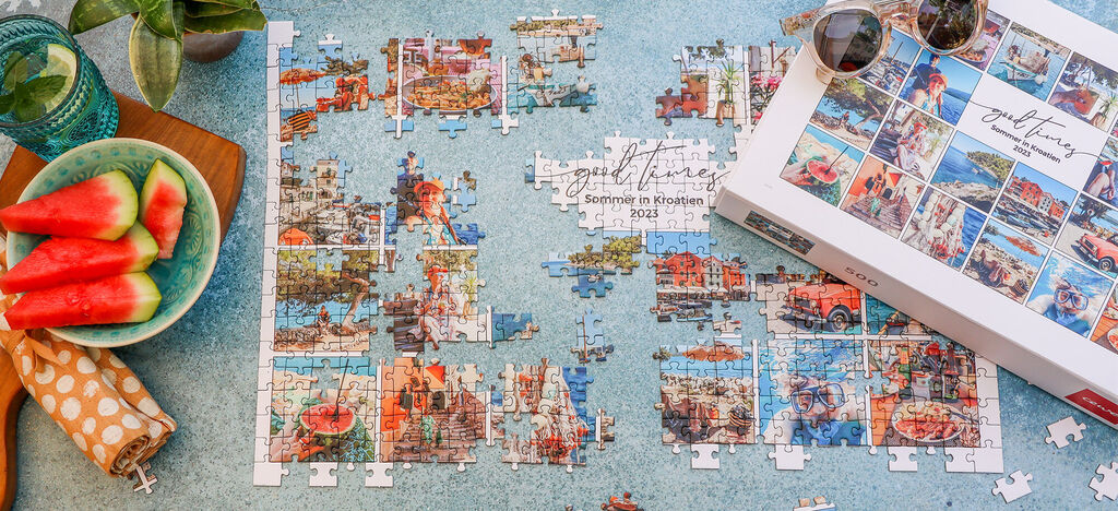 Ein fast fertiges Puzzle auf einem Tisch, umgeben von einer Schüssel Wassermelonen und einer Box mit verschiedenen Puzzlebildern.