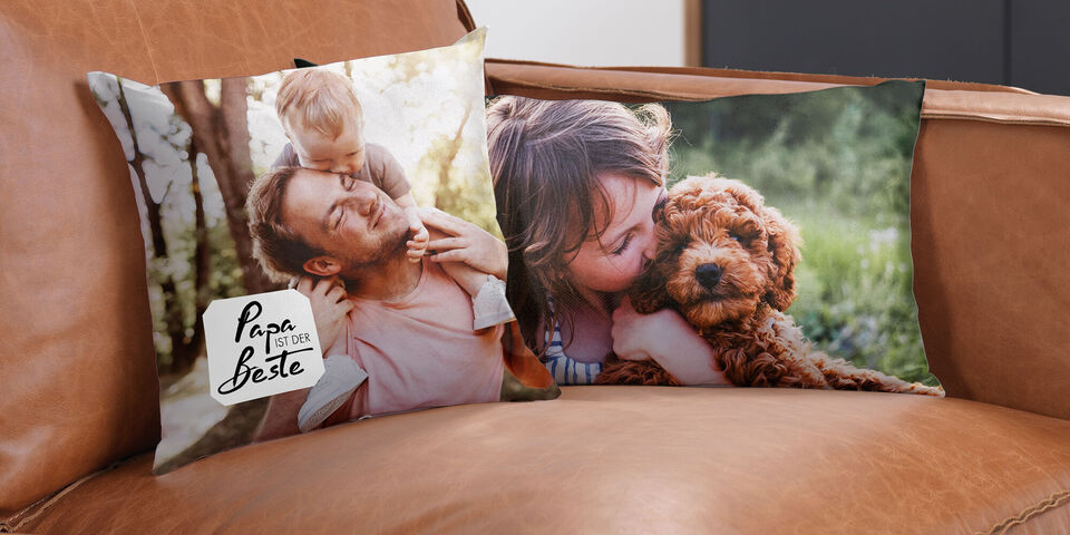 Zwei personalisierte Kissen auf einem Ledersofa. Eines zeigt einen Vater mit seinem Kind und das andere eine Frau, die einen Hund kuschelt.