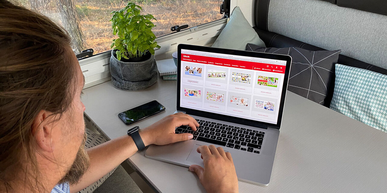 Mann sitzt am Tisch und arbeitet an einem Laptop, auf dem verschiedene Produktideen von CEWE angezeigt werden