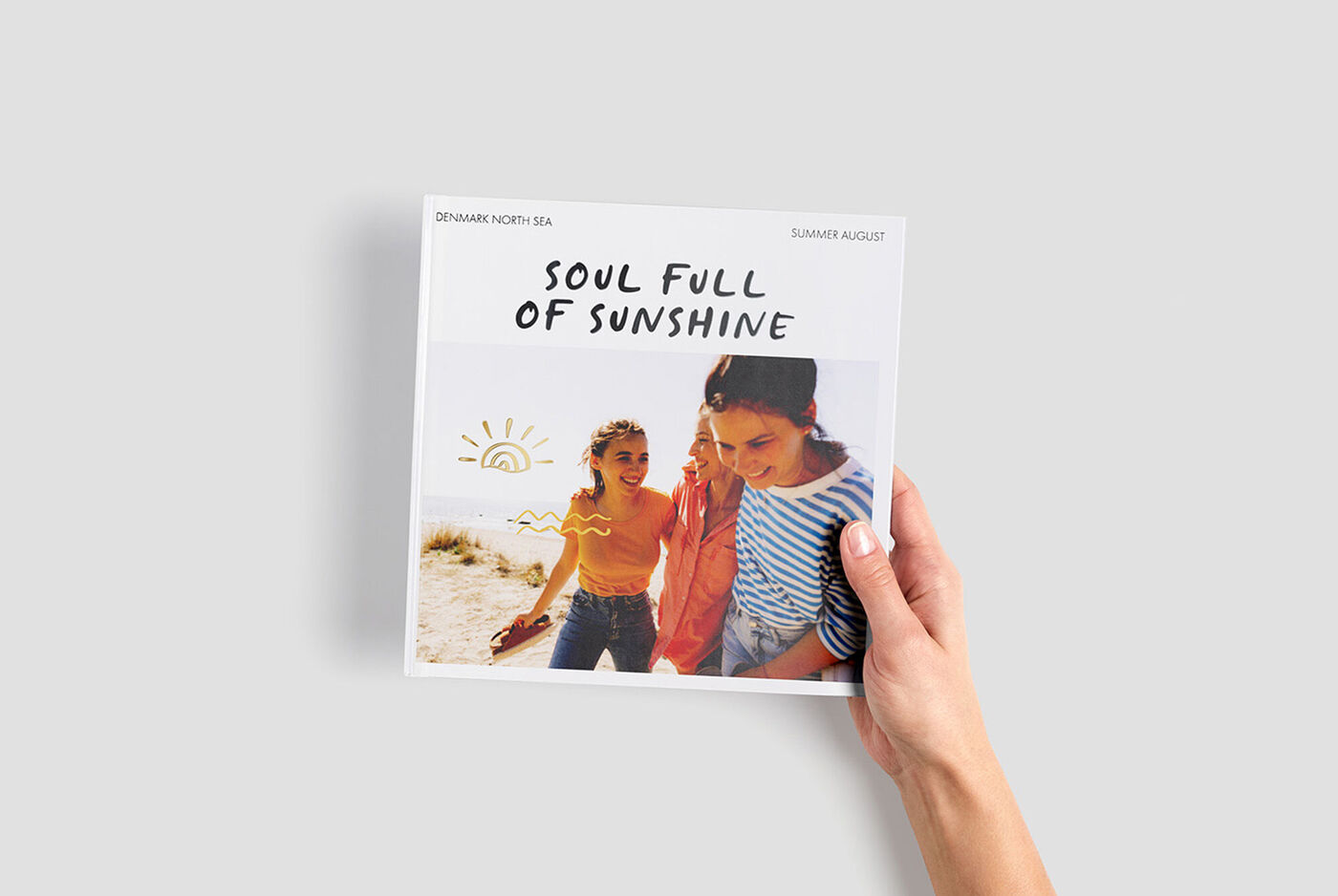Eine Person hält ein CEWE FOTOBUCH mit dem Schriftzug 'Soul Full of Sunshine' vor einem sommerlichen Hintergrund.