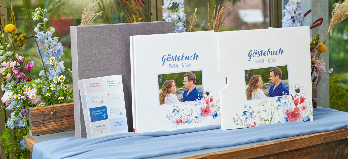 Zwei schön gestaltete Gästebücher mit floralem Design auf einem Tisch, umgeben von bunten Blumen.