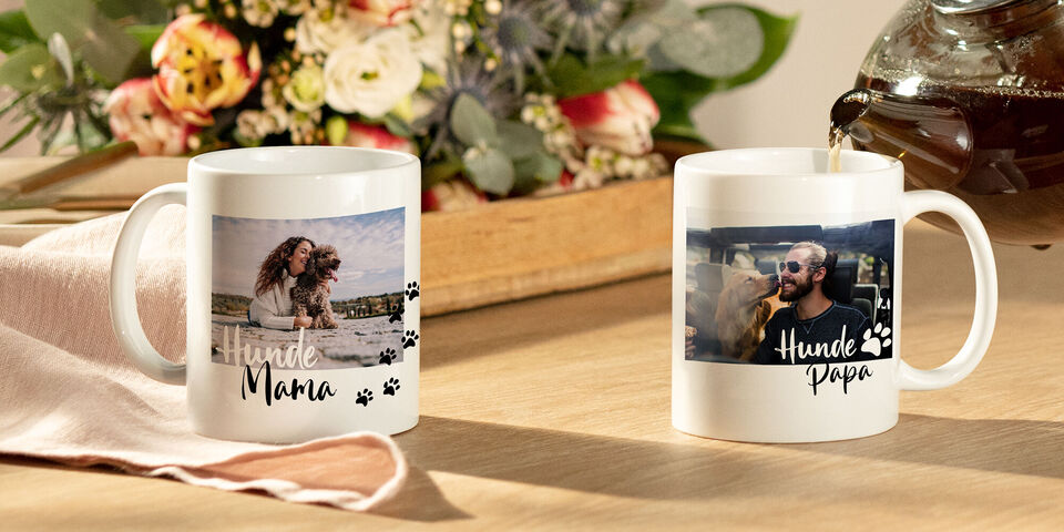 Zwei personalisierte Tassen stehen auf einem Holztisch, eine mit einem Hundebild für "Hunde Mama" und die andere für "Hunde Papa", umgeben von Blumen.