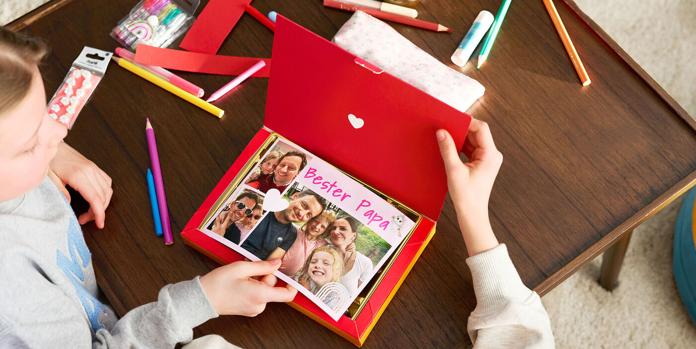 Ein Kind hält eine rote Box mit Bildern und dem Schriftzug 'Bester Papa'.