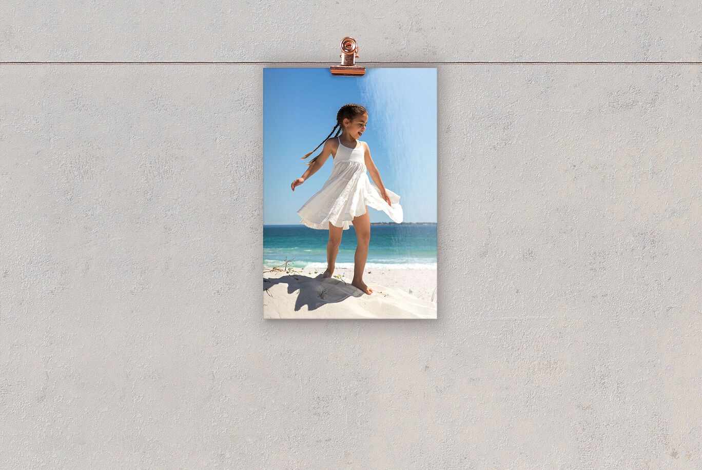 Ein fröhliches Mädchen in einem weißen Kleid steht auf einem Sandhügel am Strand. Hinter ihr ist das blaue Meer und der Himmel zu sehen.