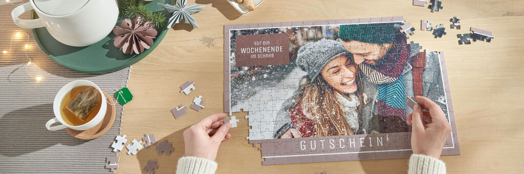 Eine Person hält einen Teil eines puzzlebasierten Gutscheins, umgeben von Puzzlestücken und einer Tasse Tee, die auf einem Tisch stehen.