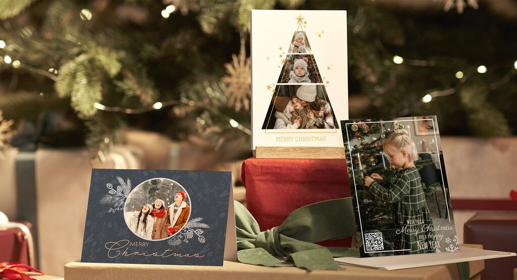 Drei festliche CEWE Grußkarten stehen liebevoll gestaltet auf Geschenken unter einem Weihnachtsbaum.