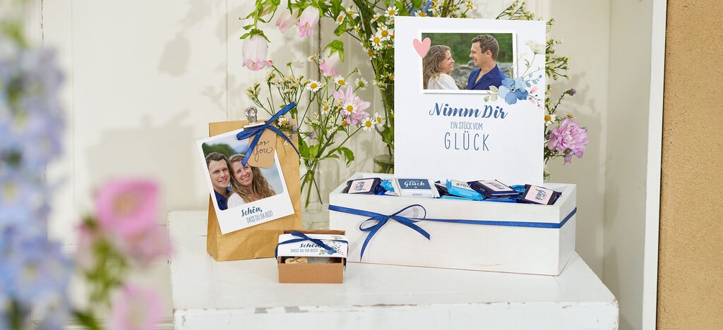 Geschenkbox und Grüße umgeben von Blumen, mit persönlichem Foto und liebevoller Botschaft.