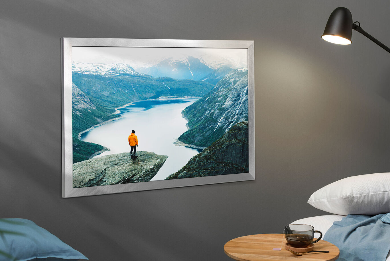 Ein Mensch mit orangefarbener Jacke steht auf einem Felsen und blickt auf einen klaren Bergsee mit schneebedeckten Bergen im Hintergrund, rahmenloses Wandbild.