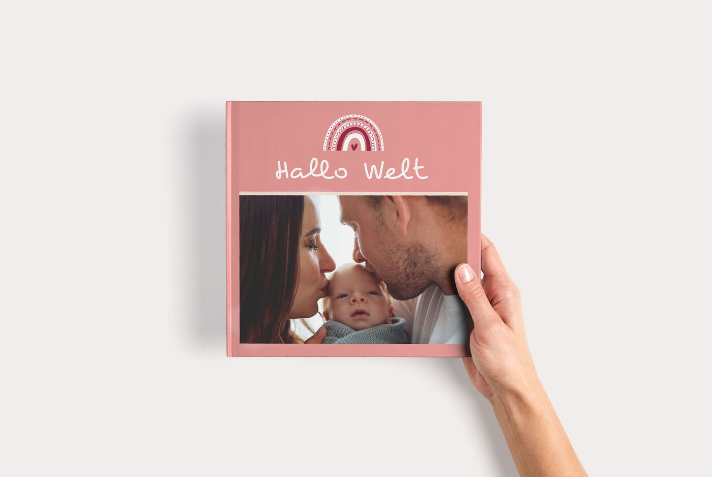 Eine Hand hält ein rosa CEWE FOTOBUCH Quadratisch zum Thema Baby mit Regenbogenillustration und einem Foto der Eltern und des Babys.