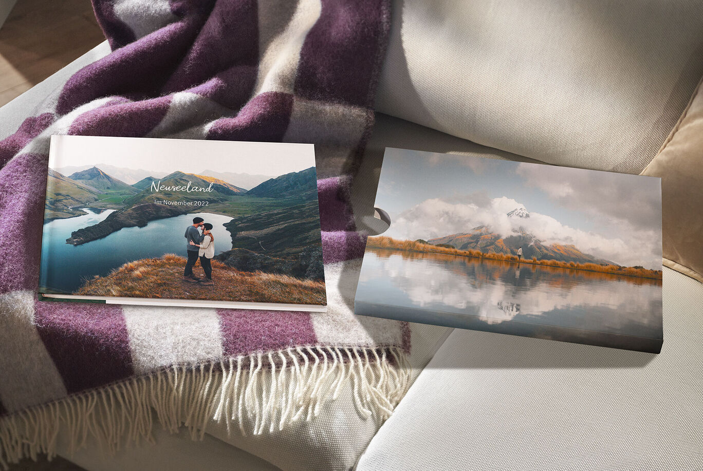 Ein CEWE FOTOBUCH liegt mit dem passenden Schuber auf einem Sofa neben einer Decke. Zu sehen sind Landschaftsmotive aus Neuseeland.