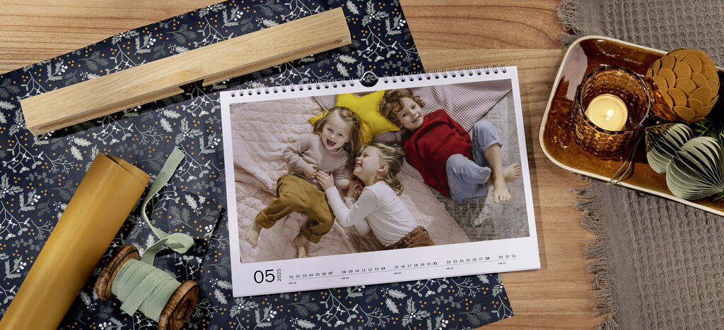 Ein kalendarisches Bild zeigt drei fröhliche Kinder, die auf einem Bett liegen und miteinander spielen. Die Kinder wirken glücklich und genießen den Moment. Der CEWE Kalender ist auf Mai eingestellt.
