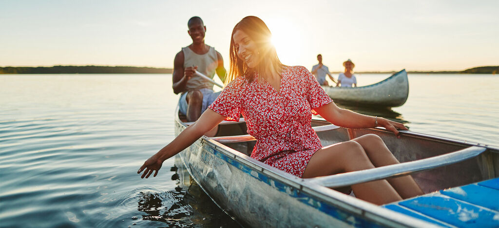 Eine Frau sitzt im Kanu und berührt das Wasser mit der Hand. Im Hintergrund sind Menschen in anderen Kanus bei Sonnenuntergang zu sehen.