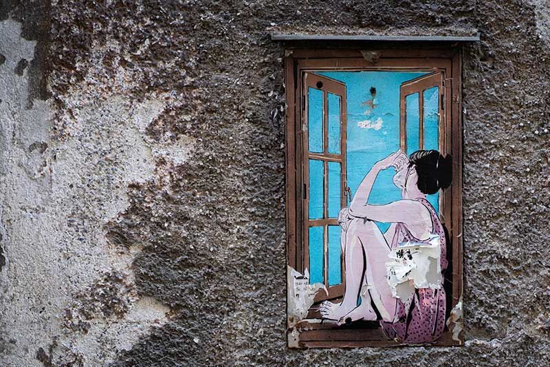 Eine Frau sitzt am Fenster, wirkt nachdenklich und schaut hinaus. Die Fenster sind geöffnet und der Himmel strahlt in sanften Blau.