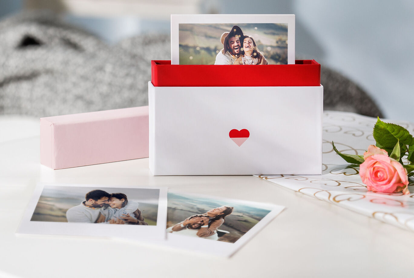 Ein liebevoll gestalteter Geschenkkarton mit Fotos. Der Karton hat einen roten Innenteil und ein Herz auf der Vorderseite. Daneben liegen zwei Fotos, die schöne Momente zeigen. Eine rosa Blume und ein designtes Blatt Papier sind in der Nähe platziert.
