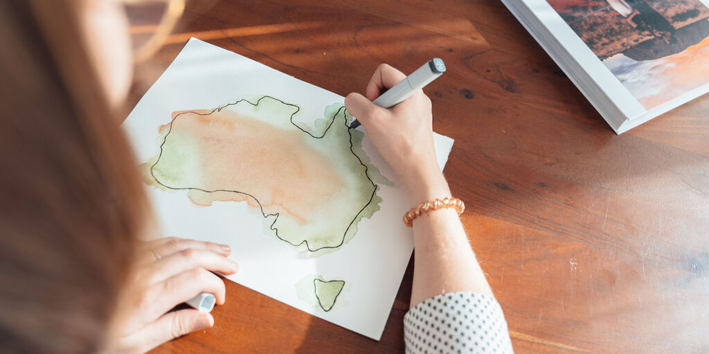Eine Person zeichnet eine farbige Australien-Karte mit einem Bleistift auf Papier.