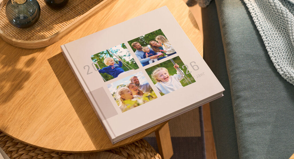 Es ist rechts ein Sofa zu sehen, auf dem eine Decke liegt. Daneben liegt ein helles CEWE FOTOBUCH mit vier Fotos auf einem runden Holztisch.