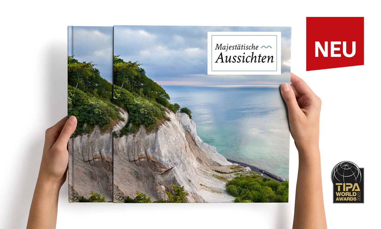 Hände halten ein CEWE FOTOBUCH mit dem Titel „Majestätische Aussichten“ und einer Küstenlandschaft auf dem Cover.