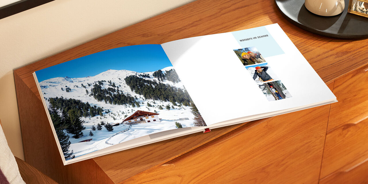 Ein offenliegendes CEWE FOTOBUCH auf einem Holzschrank, mit einer winterlichen Landschaft auf der linken Seite und Fotos auf der rechten Seite