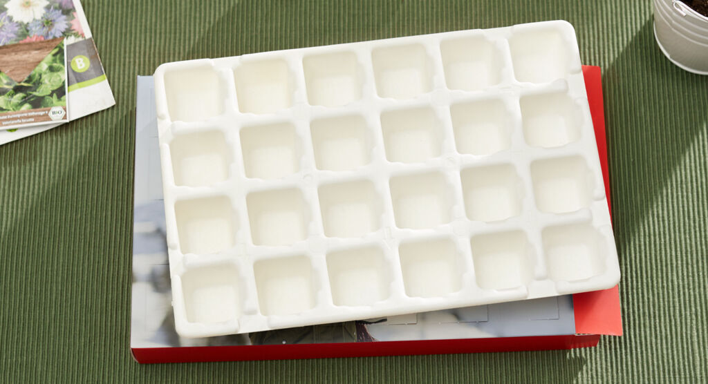 Eine weiße, platzsparende Backform mit 24 Mulden auf einem Tisch, umgeben von einer grünen Tischdecke und einer Pflanzenzeitschrift.