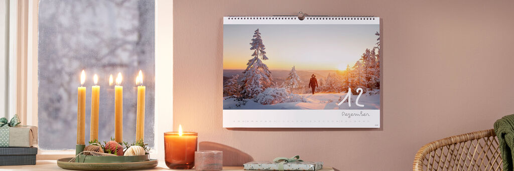 Ein CEWE Fotokalender hängt an einer Wand. Er zeigt eine wunderschöne Winterlandschaft mit Schnee und einem Sonnenuntergang. Eine Person geht in der Szene, gemütliche Kerzen und ein Dekogegenstand stehen im Vordergrund.